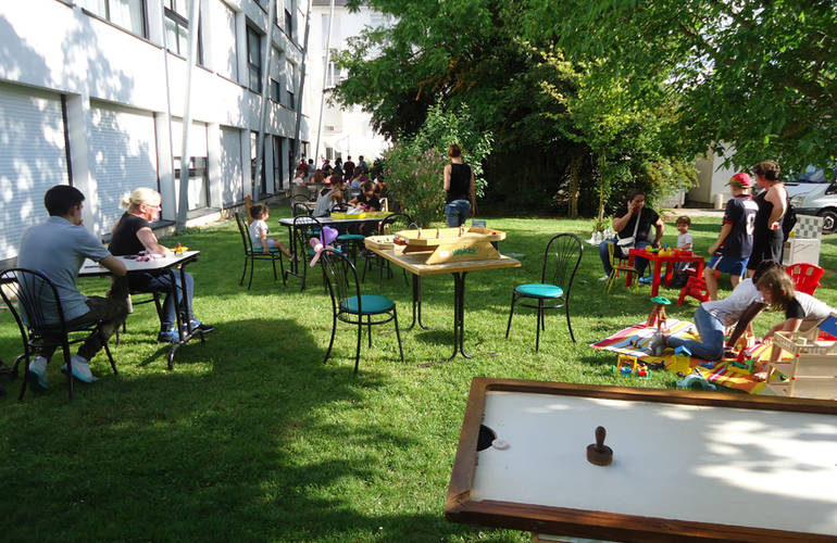 Sur la pelouse, près d'un bâtiment, des tables sont installées avec des jeux de sociétés. Des famille jouent.