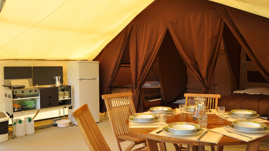 Intérieur d'une tente randonnée au camping de l’Île d'Or. En premier plan une table, au second le coin pour cuisiner et au fond, le coin chambre.