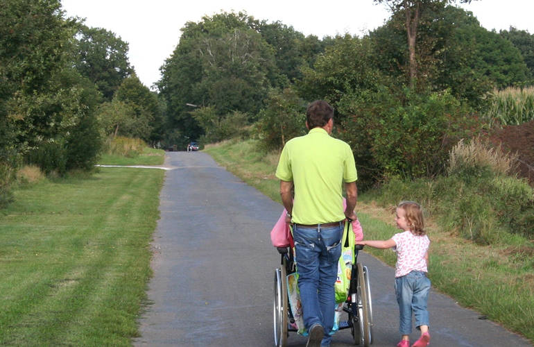 Trois personnes sur une route de campagne. Un homme pousse un fauteuil roulant, une petite fille marche à leur côté.