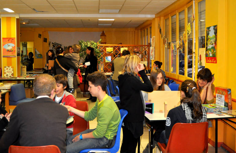 Dans une salle de nombreuses personnes jouent à des jeux assis autour de tables.