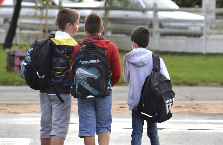 Trois enfants de dos traversent une rue pour se rendre à l'école. Ils ont tous les trois un sac à dos.