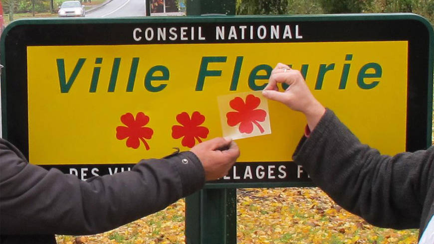 Sur un panneau de ville, 2 personnes pose l'autocollant de la troisième fleur.