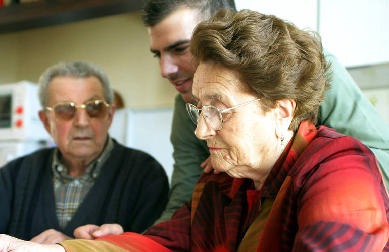 Un couple de personnes âgés se fait aider par un jeune homme. Ils sont dans une cuisine.