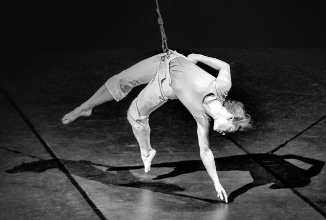 La photo est en noire et blanc. Une femme est suspendue avec grâce par un fil au niveau des hanches.