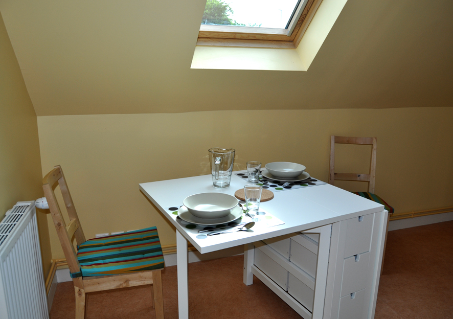 Dans une pièce en sous pente avec velux, une petite table blanche avec le couvert de mis.